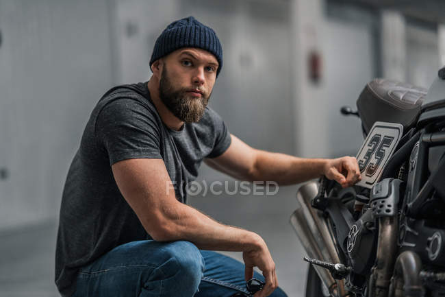 Бородатий чоловік в капелюсі піднімає брови і дивиться на камеру, сидячи біля мотоцикла на розмитому тлі сучасного гаража — стокове фото
