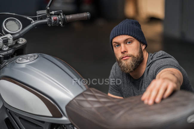 Bearded man in hat raising eyebrow and looking at camera while sitting near motorcycle on blurred background of contemporary garage — Stock Photo