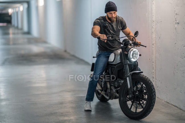 Corpo inteiro barbudo cara em roupas casuais sentado na motocicleta e olhando para longe no corredor da garagem moderna — Fotografia de Stock