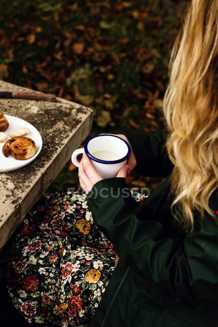 Зверху врожаю жінка в теплому повсякденному одязі п'є молоко і їсть смачну домашню випічку, сидячи за сірим кам'яним столом з букетом свіжих жовтих диких квітів в осінньому саду — стокове фото