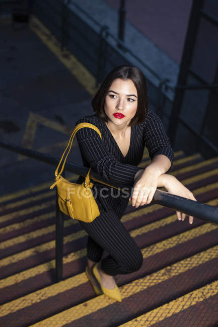 Modische Frau in schwarzem Kleid mit rotem Lippenstift und gelber Tasche lehnt in der Abenddämmerung am Treppengeländer einer Stadtstraße — Stockfoto