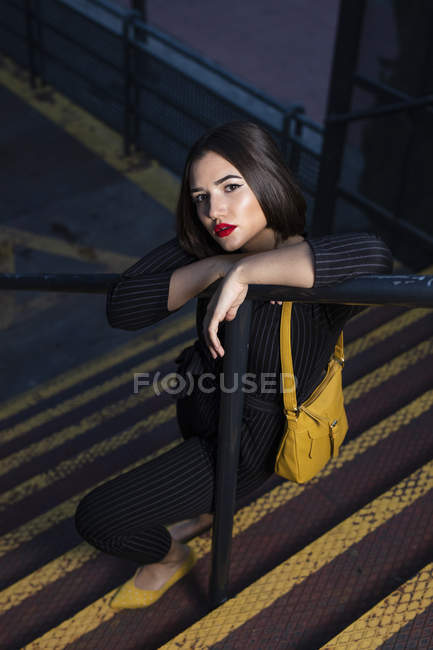 Fashionable woman in black dress with red lipstick and yellow small bag getting into peaky cap leaning at staircase handrail in a city street on dusk — Stock Photo