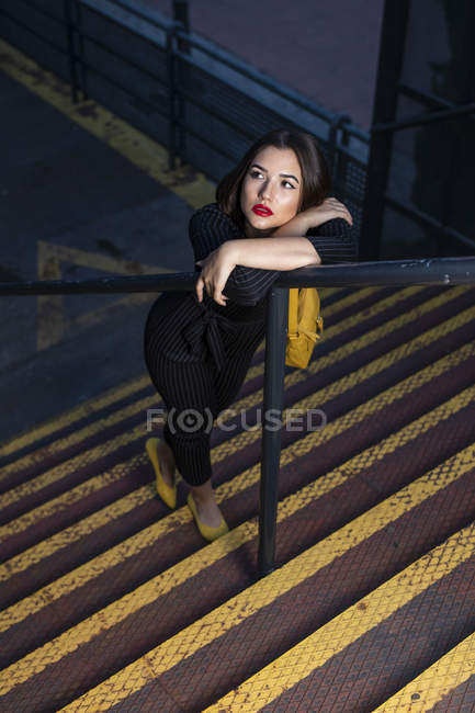 Donna alla moda in abito nero con rossetto rosso e borsa gialla piccola appoggiata al corrimano scala in una strada cittadina al tramonto — Foto stock