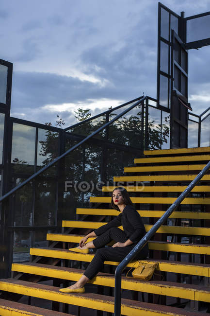 Donna alla moda in abito nero con rossetto rosso e borsa gialla piccola seduta al corrimano scala in una strada cittadina al tramonto — Foto stock