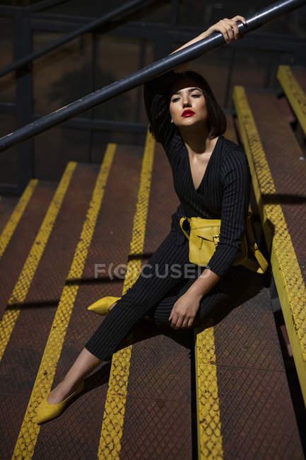 Donna alla moda in abito nero con rossetto rosso e borsa gialla piccola seduta al corrimano scala in una strada cittadina al tramonto — Foto stock