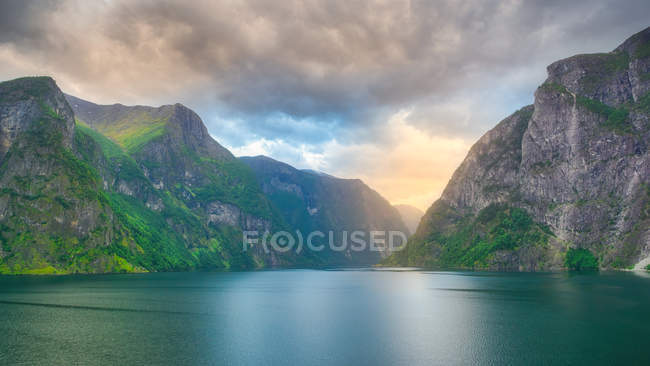 Потрясающий пейзаж на зеленой воде, отражающей облачное небо, омывающий скалистые горы зеленым деревом и травой в Норвегии — стоковое фото