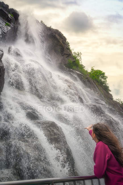 Rückansicht einer langhaarigen Frau, die den schnellen Gebirgsfluss beobachtet, der in Norwegen steinige, mit grünem Gras bedeckte Hügel hinunterfließt — Stockfoto