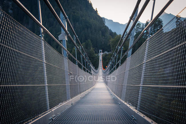 Вечером в Австрии над оврагом в зеленых горах расположен современный металлический подвесной мост с высокими перилами — стоковое фото
