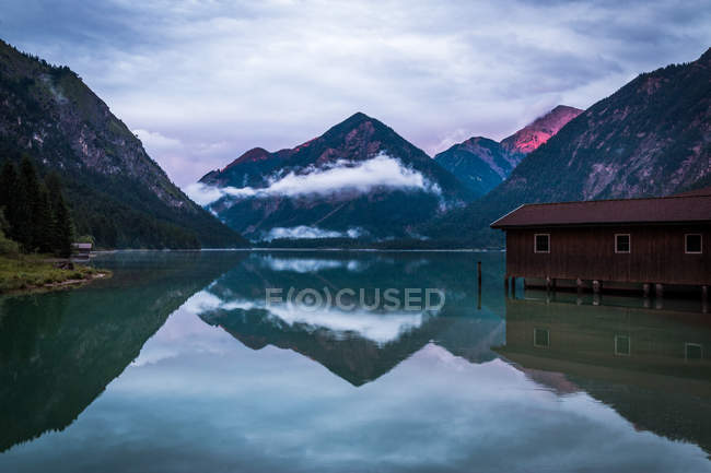 Shabby capannone per le barche situate vicino all'acqua tranquilla dello stagno vicino cresta di montagna il giorno nuvoloso in Austria — Foto stock