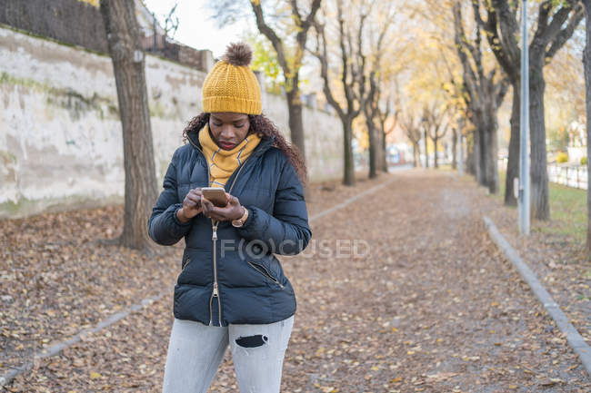 Femme afro-américaine au chapeau jaune et veste chaude en utilisant un smartphone sur la route avec des feuilles d'automne dans le parc — Photo de stock