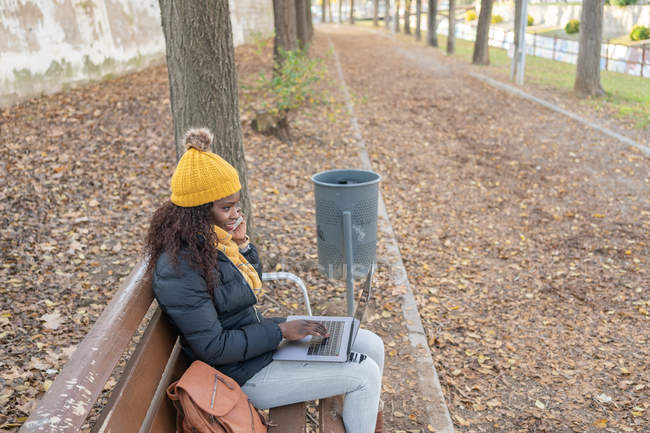 Seitenansicht der fröhlichen Mode afrikanisch-amerikanische Frau mit gelbem Hut und warmer Jacke arbeitet auf Laptop bequem auf Holzbank im Herbst Park sitzen — Stockfoto