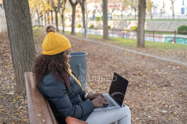 Side view of cheerful fashion African American woman in yellow hat and warm jacket working on laptop comfortably sitting on wooden bench in autumn park — Stock Photo