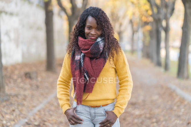 Чарівна жінка з Африки в зимовому шарфі з руками в кишені на дорозі з осіннім листям в парку — стокове фото