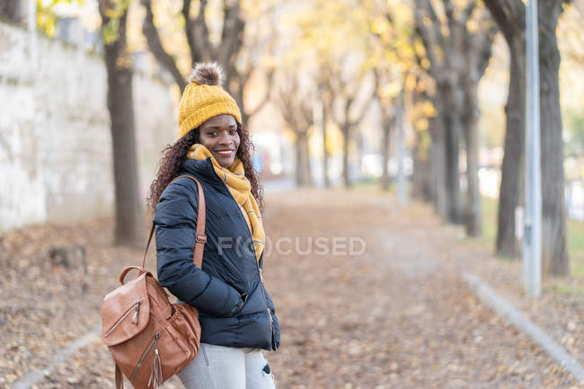 Enthusiastic trendy African American woman in yellow hat and warm jacket with leather backpack and hands in pocket looking at camera with smile on road autumn park — Stock Photo