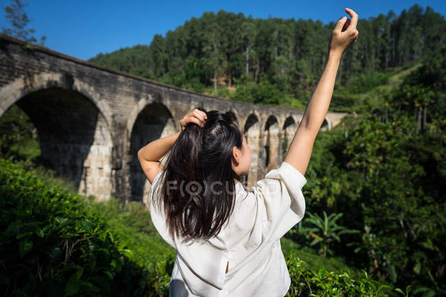 Visão traseira da mulher levantando a mão para cima desfrutando de uma vista incrível da velha ponte cercada por floresta verde em Nine Arches Bridge, Ella, Sri Lanka — Fotografia de Stock