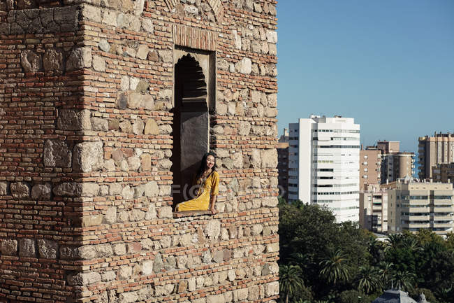 Asiatische Reisende lächelt, während sie im Fenster des Alcazaba-Gebäudes an einem sonnigen Tag in Malaga, Spanien sitzt — Stockfoto