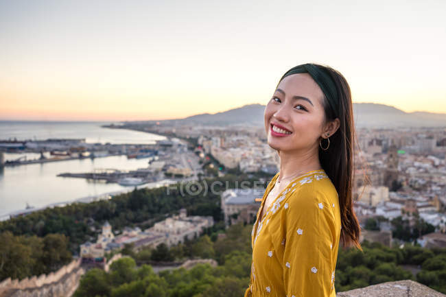 Mulher asiática sorrindo e olhando para a câmera enquanto visita Castillo Gibralfaro no fundo borrado da cidade costeira e do céu do pôr-do-sol em Málaga, Espanha — Fotografia de Stock