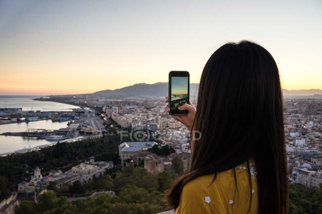 Vue arrière de la femme utilisant un smartphone pour prendre des photos de la ville côtière et du ciel sans nuages au coucher du soleil lors de la visite de Castillo Gibralfaro à Malaga, Espagne — Photo de stock