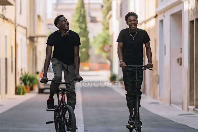 Un jeune Afro-américain gai regarde une caméra et un scooter électrique tandis qu'un ami noir regarde vers le haut et conduit une bicyclette dans la rue — Photo de stock