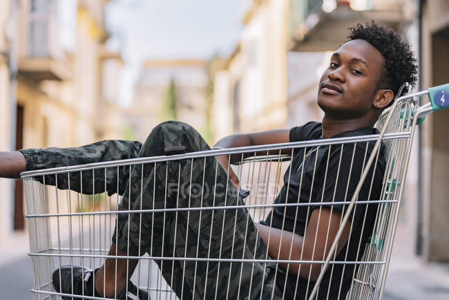 Side view of pensive youthful African American male teenager in casual clothes looking at camera while sitting in metal shopping trolley in street. — Photo de stock