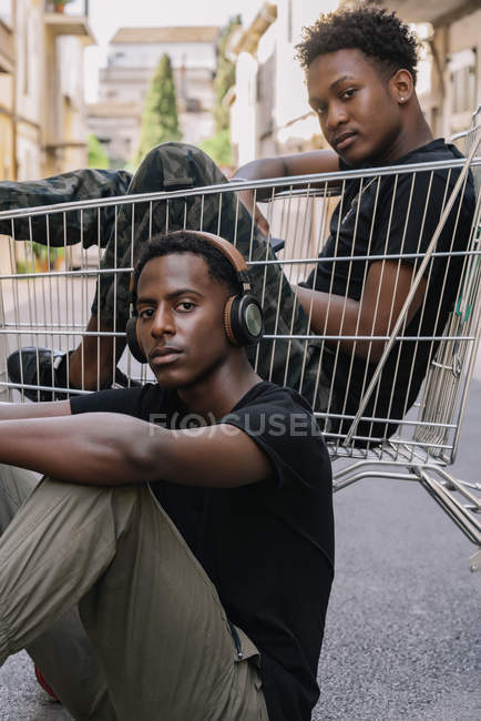 Feliz hombre afroamericano con ropa casual sentado en carro con un amigo masculino negro en los auriculares. - foto de stock
