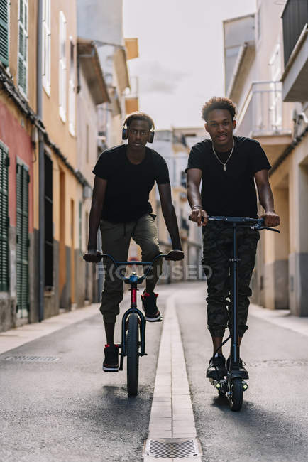 Cheerful young African American man riding electric scooter while black male is driving bicycle in street looking at camera — Stock Photo