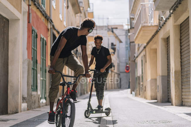 Un jeune Afro-Américain se promène dans un scooter électrique tandis qu'un ami noir conduit une bicyclette dans la rue — Photo de stock