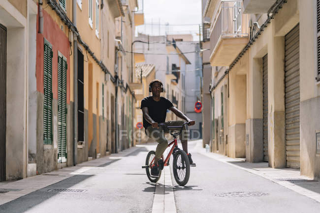 Focused youthful African American male teenager in headphones riding BMX bike in sunshine in neighborhood — Stock Photo