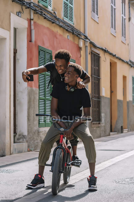 Счастливые молодые афроамериканские подростки делают селфи на мобильный телефон, сидя вместе на велосипеде Bmx на улице — стоковое фото