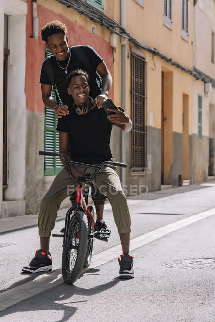 Happy youthful African American male teenagers taking selfie on cellphone while sitting on BMX bike together in street — Stock Photo
