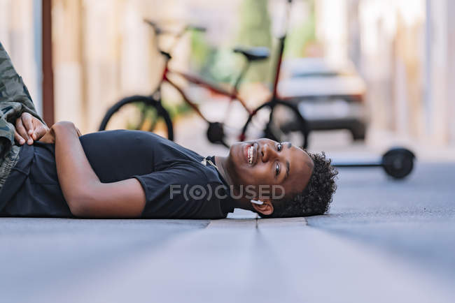 Side view of happy youthful African American man lying on asphalt road in street looking at camera. — Photo de stock
