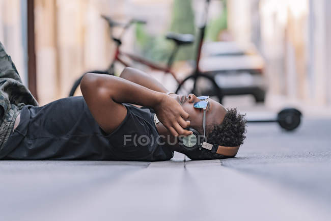 Side view of serious youthful African American man in sunglasses and headphones listening to music while lying on asphalt road in street — Stock Photo