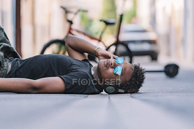 Vista lateral del joven y serio afroamericano en gafas de sol y auriculares que escuchan música mientras se encuentra en la carretera asfaltada de la calle. - foto de stock