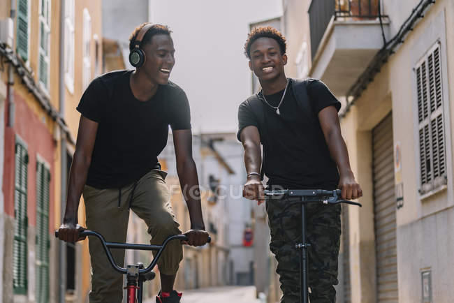Веселый молодой афроамериканец смотрит в камеру и ездит на электрическом скутере, в то время как его друг-чернокожий мужчина смотрит вверх и ездит на велосипеде по улице — стоковое фото