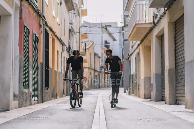 Веселий молодий афроамериканець їде на електричному скутері, а чорний чоловік їздить на велосипеді на вулиці. — стокове фото