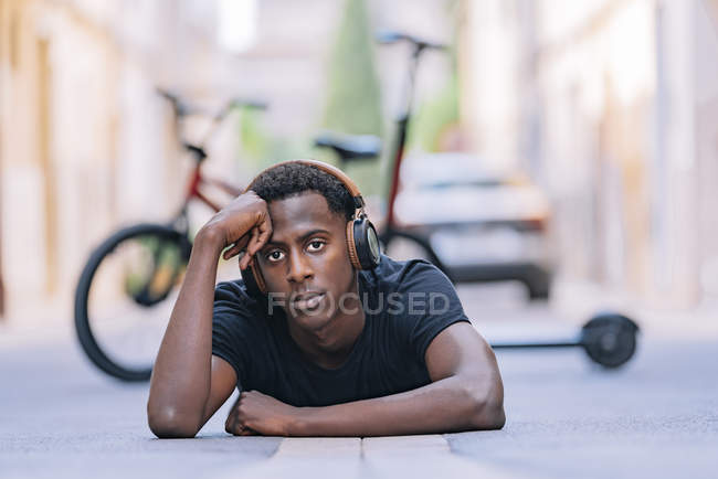 Concentrated youthful African American man wearing headphones listening to music while lying on asphalt road in street — Stock Photo