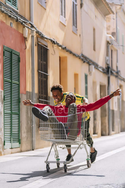 Happy carefree young African American friends in casual clothes riding around in shopping trolley in street — Stock Photo