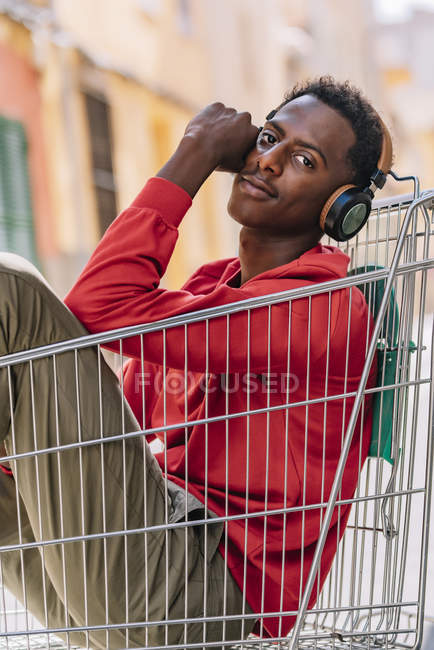 Вид сбоку на застенчивого юношу-афроамериканца в повседневной одежде, смотрящего в камеру, сидя в металлическом торговом тролли на улице — стоковое фото