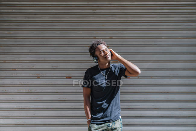 Stylish modern African American man with closed eyes and headphones listening to music at striped wall in street — Stock Photo