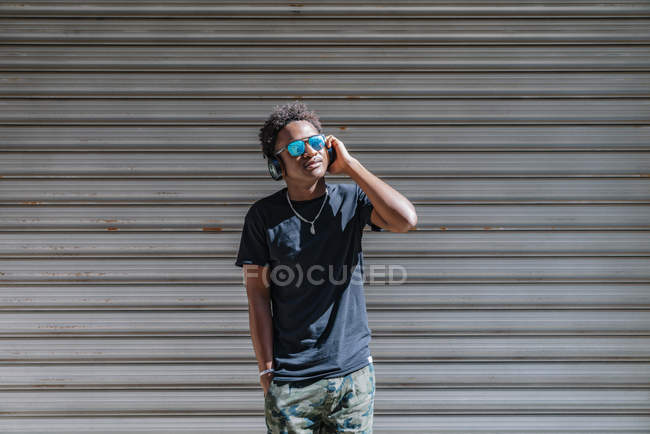 Homme moderne afro-américain élégant portant des lunettes de soleil et des écouteurs qui écoutent de la musique au mur rayé dans la rue — Photo de stock