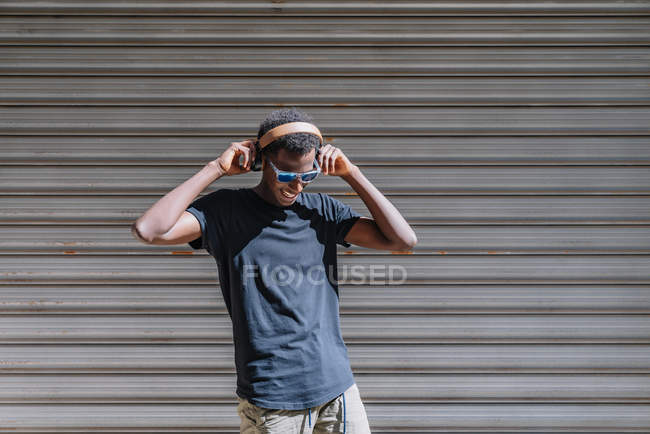 Stylish modern African American man in sunglasses and headphones listening to music at striped wall in street — Stock Photo