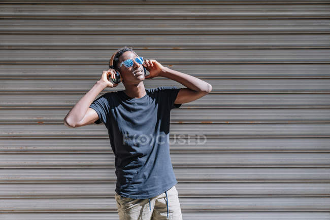 Elegante homem americano Africano moderno em óculos de sol e fones de ouvido ouvir música na parede listrada na rua — Fotografia de Stock