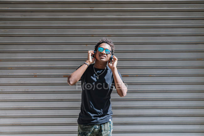 Estilo hombre afroamericano moderno en gafas de sol y audífonos que escucha música en la pared rayada de la calle. - foto de stock