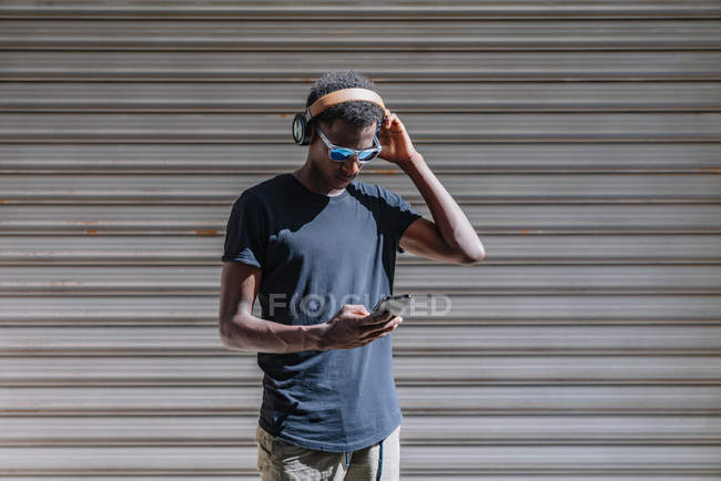 Stylish modern African American man in sunglasses and headphones listening to music on the mobile phone at striped wall in street — Stock Photo
