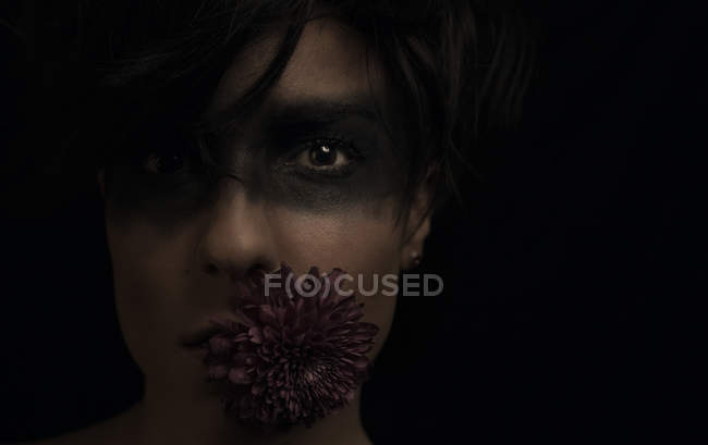 Crop thoughtful young dark haired female model with black color around eyes and flower in mouth looking at camera in studio on black background — Stock Photo