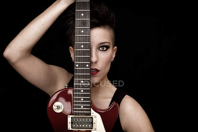 Femme avec maquillage lumineux et cheveux courts foncés regardant la caméra et couvrant la moitié du visage avec guitare électrique en studio sur fond noir — Photo de stock
