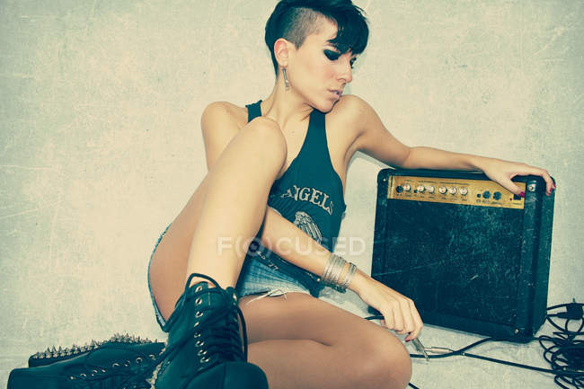 Nachdenkliche junge Frau in Rockerkleidung und mit moderner Frisur, die mit Retro-Gitarren-Combo-Verstärker im Studio auf dem Boden sitzt — Stockfoto