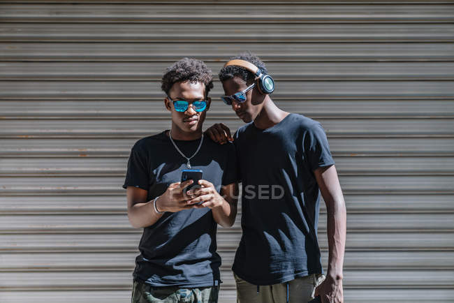 Крутые молодые афроамериканские подростки в солнцезащитных очках, фотографирующиеся с мобильным телефоном, стоя на солнце на улице — стоковое фото