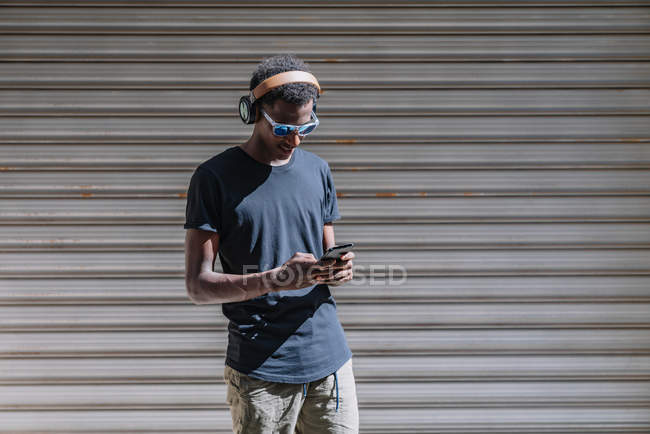 Estilizado hombre afroamericano moderno en gafas de sol y audífonos que escucha música en el teléfono móvil en la pared rayada de la calle. - foto de stock