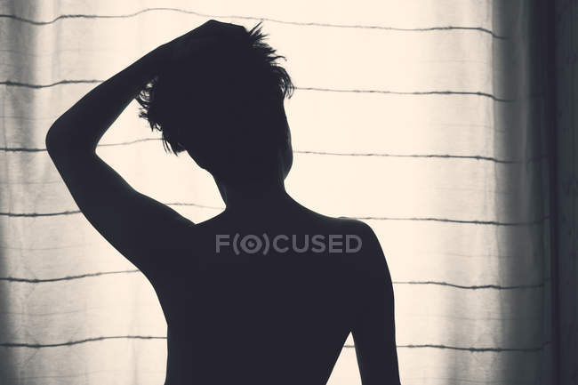 Вид сзади на неузнаваемый силуэт женщины с короткими волосами, поднимающейся рукой вверх и стоящей перед окном с занавесом — стоковое фото